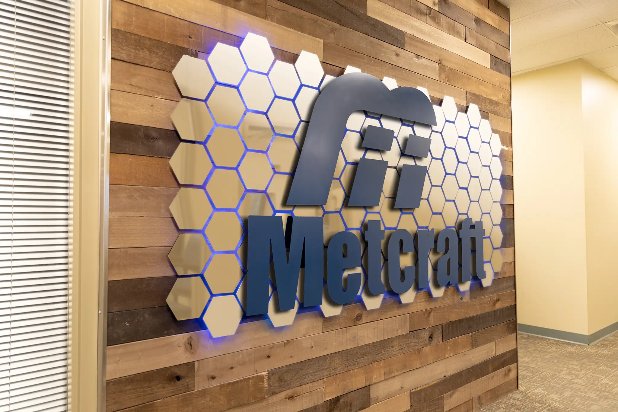 A metal sign with repeating, shiny hexagons behind a blue Metcraft logo, installed on a wood wall in a hallway.